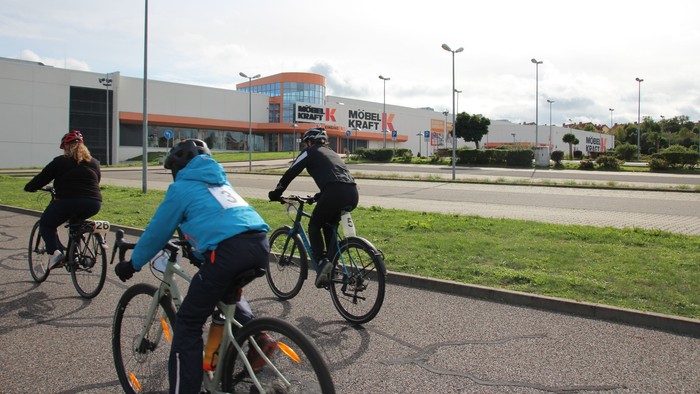 Drei Radfahrer fahren auf einem Radweg an einem Einkaufszentrum vorbei.