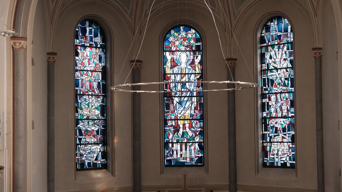 Bunte Glasfenster und ein hängender Kronleuchter in einem prächtigen, gewölbten Kircheninnenraum.