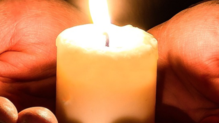 Hænder, der forsigtigt holder et tændt stearinlys mod en mørk baggrund, som kaster et varmt, glødende lys.