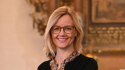 En kvinde i sort jakke og briller står ved et podium i en historisk lokalitet.