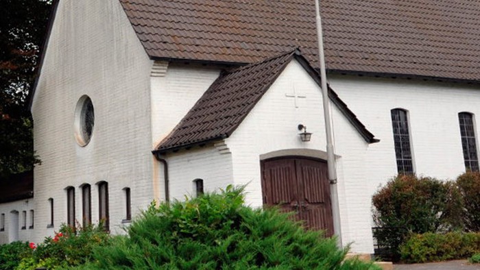 Weiße Kirche mit hohem Turm und braunem Dach, umgeben von Bäumen und Büschen