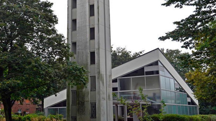 Das Bild zeigt eine moderne Kirche mit hohem Turm und Glasfront, umgeben von Bäumen.