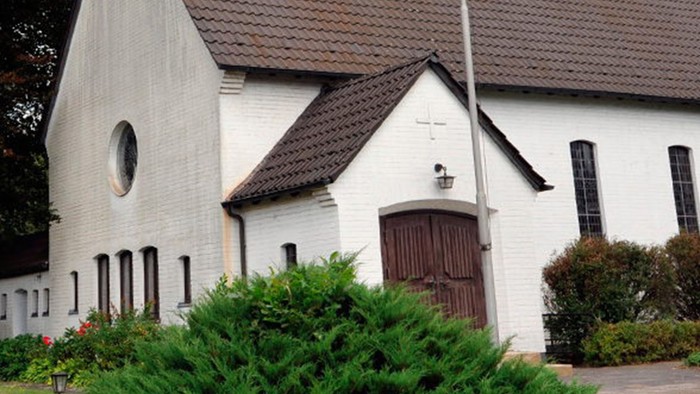 Weiße Kirche mit hohem Turm und braunem Dach, umgeben von Bäumen und Büschen