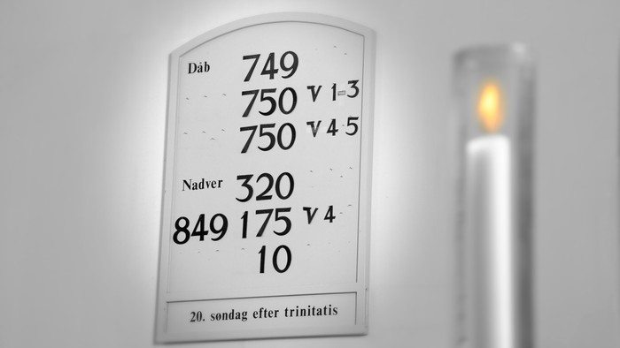 Et tændt lys ved siden af en kirkegudstjeneste-salmebog med nummererede læsninger og datoer på dansk.