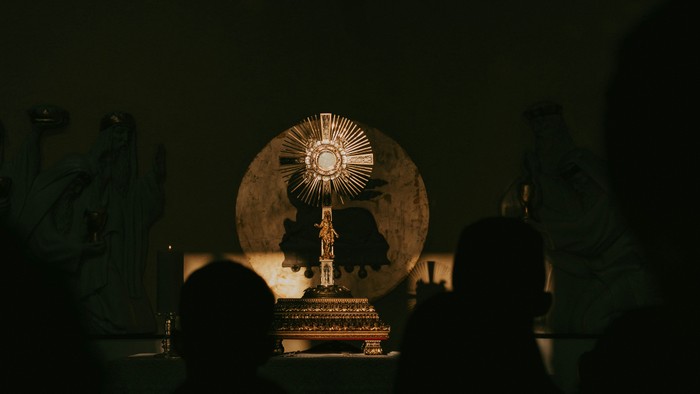 Zwei Personen beten vor einem beleuchteten Kreuz in einer dunklen Kirche.
