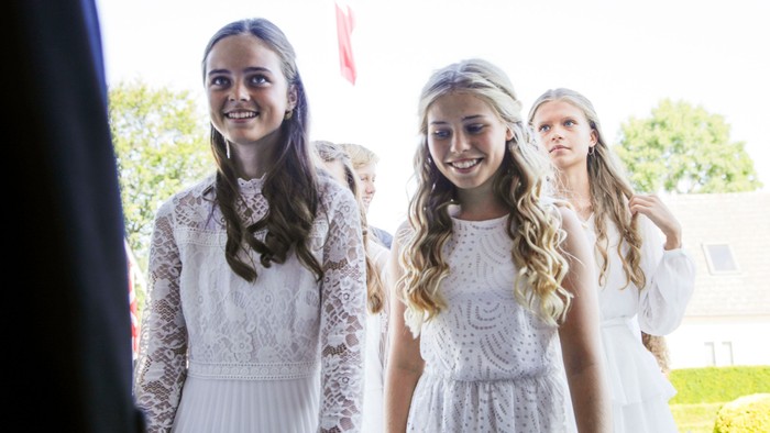 Tre unge piger i hvide spidsbløse kleder smiler og står sammen ude.