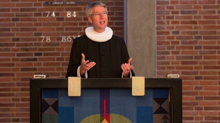 En kvinde står bag et podie på et scene med mur af røde mursten.