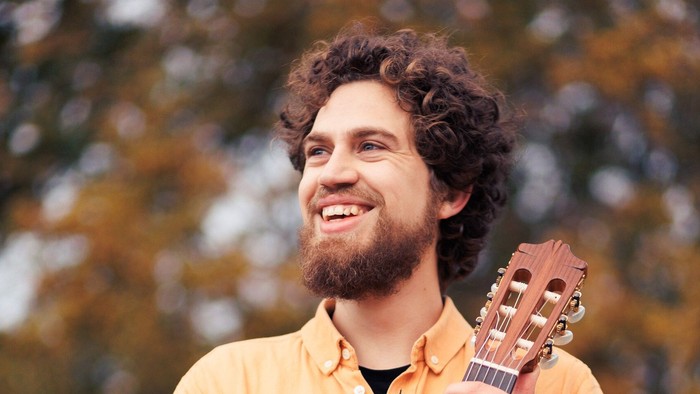 En glad mand i en gul skjorte holder og smiler, mens han spiller på en lysebrun guitar udendørs.