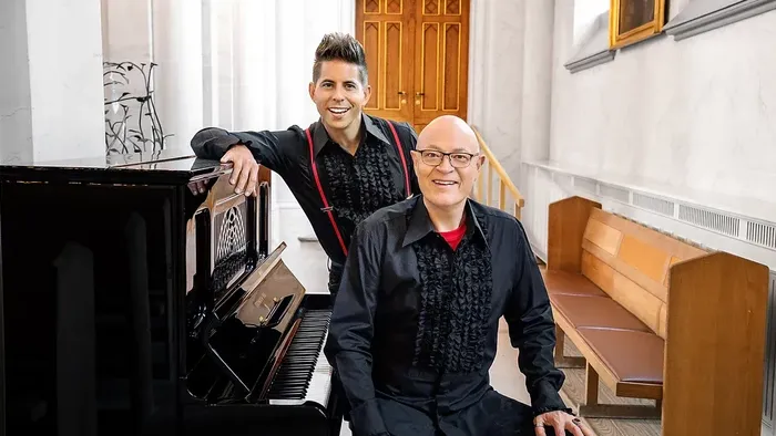 To mænd poser sammen nær et flygel i en kirke, den ene står op, og den anden sidder ned.