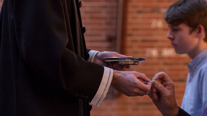 En mand i mørkt tøj holder en mobiltelefon, mens en ung dreng i blå tøj ser på.