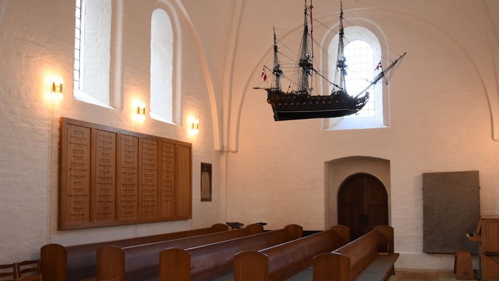 En skibsmodel hænger i et historisk kirkerum med træbænke.