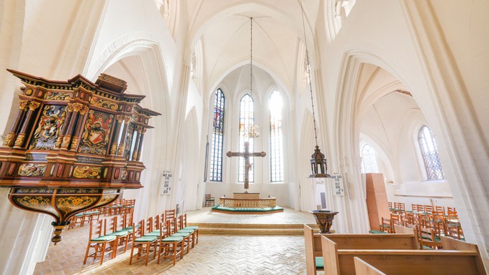Elegant kirkeinteriør med udsmykket prædikestol, træbænke og buede farvede glasvinduer under blødt lys.