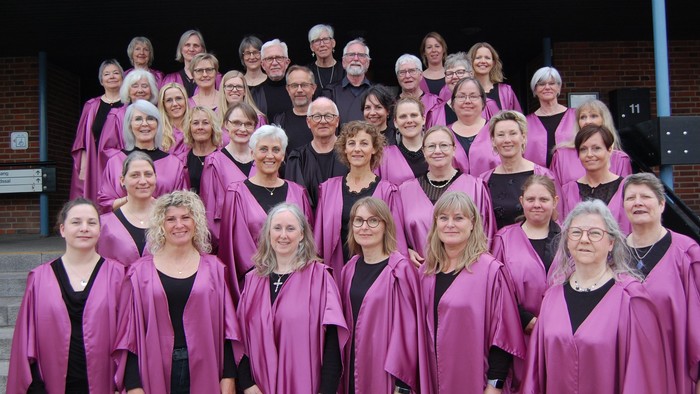 En stor kor, der består af mange kvinder, står sammen i rækker og smiler til kameraet. De er alle iført rosa kapper og sorte skjorter.
