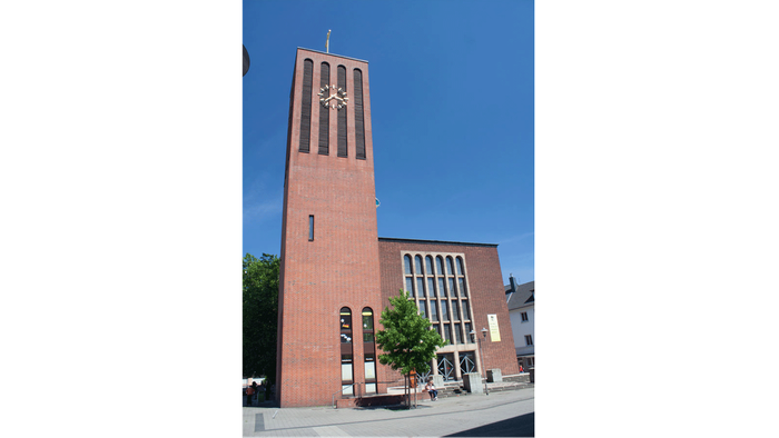 Backsteinkirche mit hohem Turm und Uhr, umgeben von Bäumen und einer Straße.