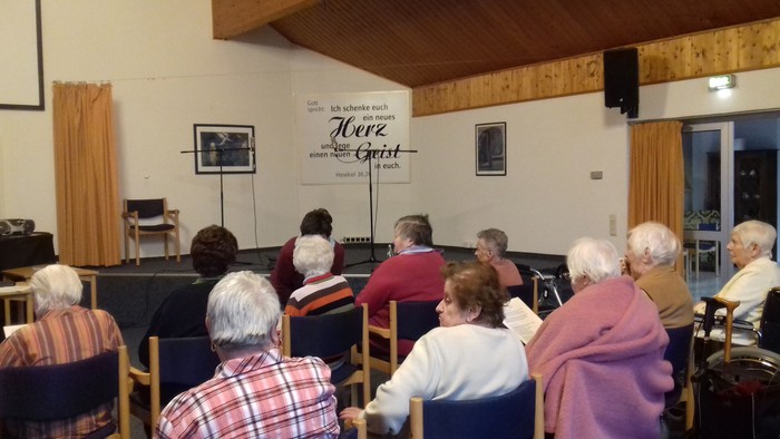 Eine Gruppe älterer Menschen sitzt in einem Raum und schaut einem Vortrag zu.