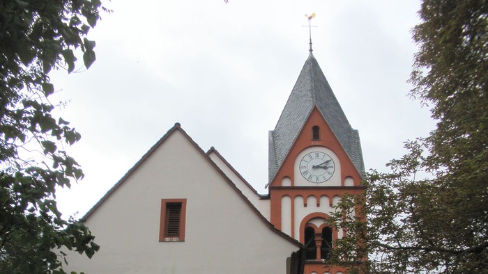 Kleine Kirche mit hohem Turm und Uhr, umgeben von Bäumen und Büschen