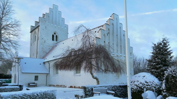 En snedækket kirke med tårn og en træ i forgrunden