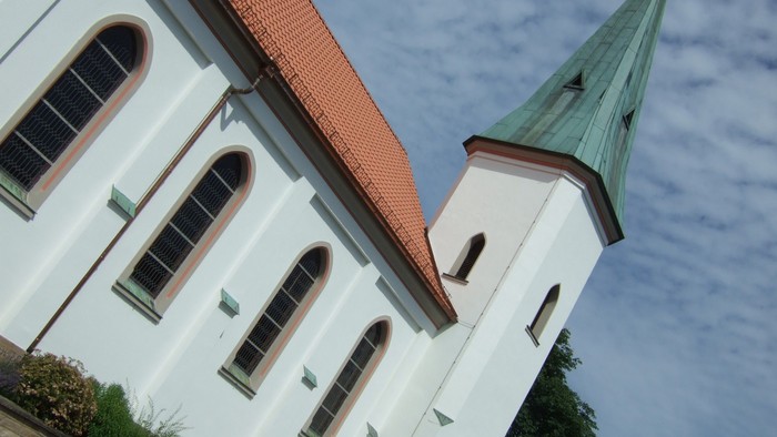 Eine weiße Kirche mit Bogenfenstern und einem grün gedeckten Kirchturm vor klarem Himmel.
