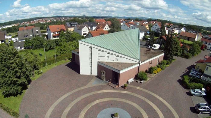Luftaufnahme einer modernen Kirche, umgeben von Wohnhäusern und einem kreisförmigen gepflasterten Platz.