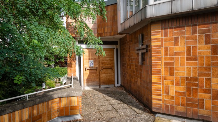 Das Bild zeigt einen Gehweg neben einem Gebäude mit Ziegelmauerwerk und Bäumen.