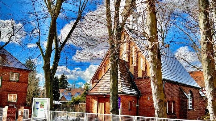 Ein rotes Backsteingebäude mit spitzem Dach und einem Kreuz darauf, umgeben von kahlen Bäumen und einem blauen Himmel.