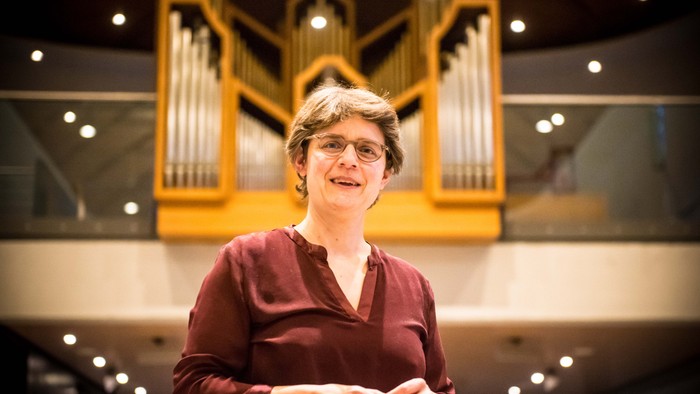 Eine Person steht lächelnd vor einer großen, beleuchteten Pfeifenorgel in einem Konzertsaal.