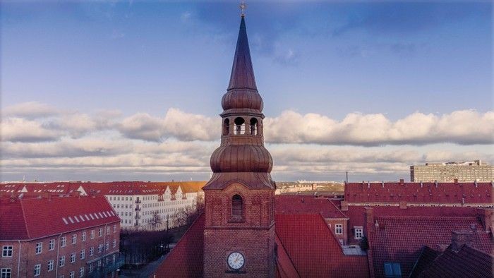 Historisk kirkespire, der rejser sig over en by med rødstens tage under en delvist overskyet himmel.