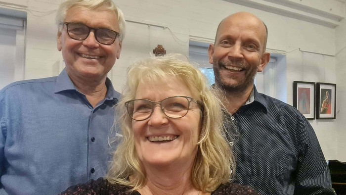 Tre glade voksne, der smiler sammen i et lyst indendørs miljø.