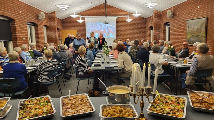 En gruppe af mennesker sidder ved bord i en lokal og lytter til en foredragsholder. Der er mad på bordene foran.
