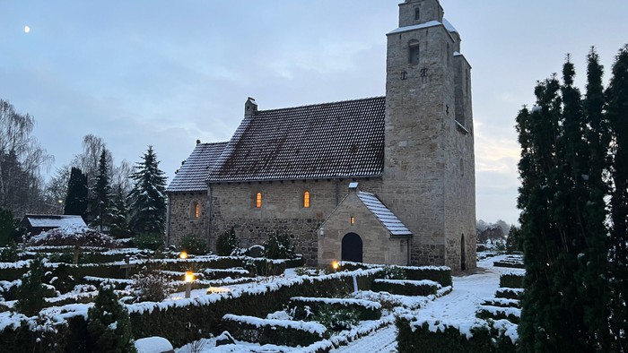En snede kirke med tårn og lysende vinduer omgivet af sne og træer