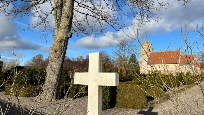 Et hvidt kors står nær et træ og en bygning med et rødt tag under en blå himmel.