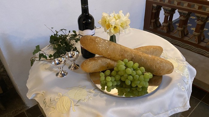 En hvid duk dekker bordet med brød, druer, vinflaske og blomster.