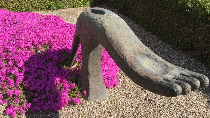 En stor bronzeskulptur af en fod, der stikker op af jorden mellem levende lyserøde blomster. Ved fostergraven
