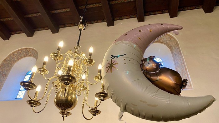 En stor blå ballong i form af en halvmåne hænger fra loftet i et rum med håndværkede træbalker og en storslåen kandelier.