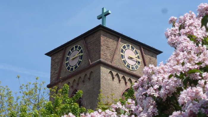 Ein Kirchturm mit Uhr und Kreuz, umgeben von blühenden Bäumen und einem klaren blauen Himmel