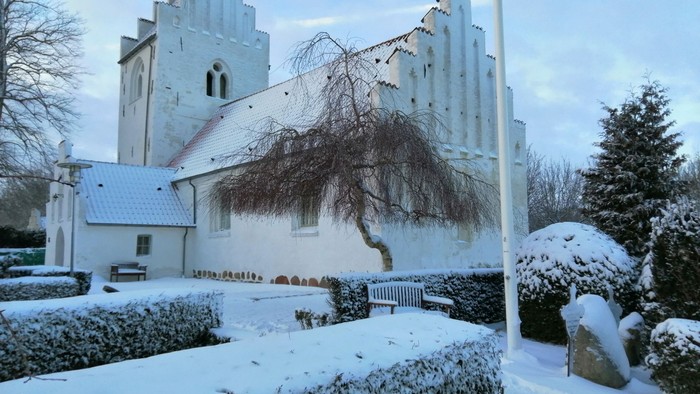 En snedækket kirke med tårn og en træ i forgrunden