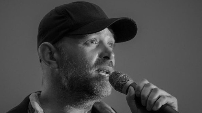 En mand med kasket taler lidenskabeligt ind i en mikrofon, fanget i et stemningsfuldt sort-hvidt portræt.