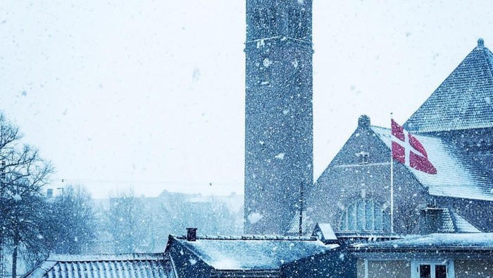 Et vinterlandskab dækket af sne med et højt klokketårn og sneklædte bygninger med et dansk flag, der vajer.