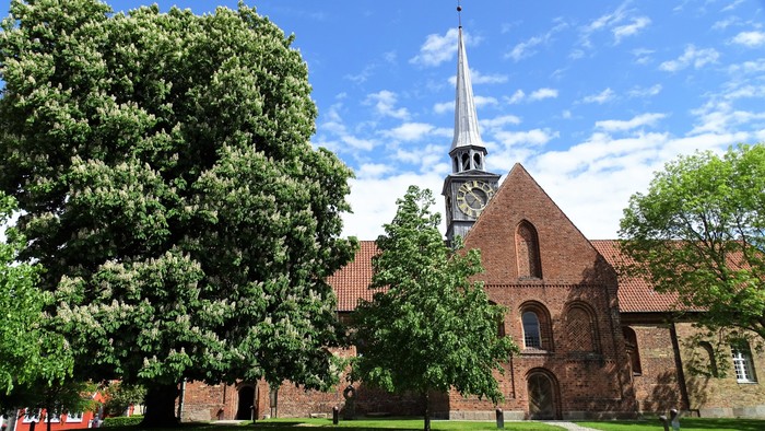 En rød murstenskirke med tårn og klokke, omgivet af grønne træer og en græsplæne