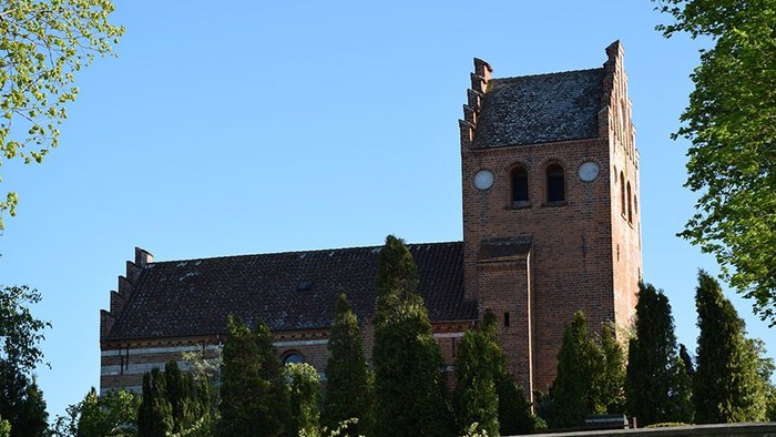 En gammel kirke med tårn og rundbuefænestre, omgivet af træer og buske