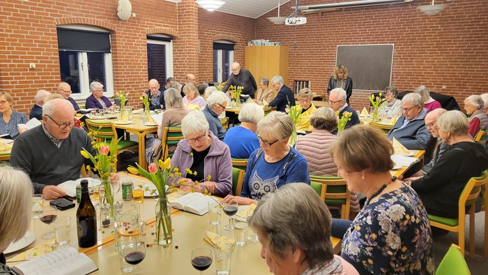 En gruppe ældre samles til et fællesspisning i et rum med mur af røde teglsten.