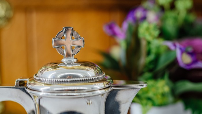 En sølv tekanne med kors på lokket og blomster i baggrunden