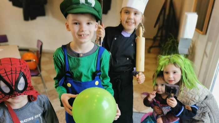 Fire børn i fantasikostumer, et børn med grøn hat og ballong, et børn med spiderman-masker, et børn med køkkentøj og et børn med grøn hår og tute.
