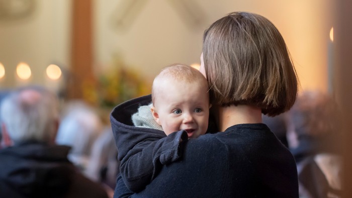Eine Frau hält ein Baby während eines feierlichen Gottesdienstes in einer Kirche mit einem Kreuz und religiösen Symbolen im Hintergrund.