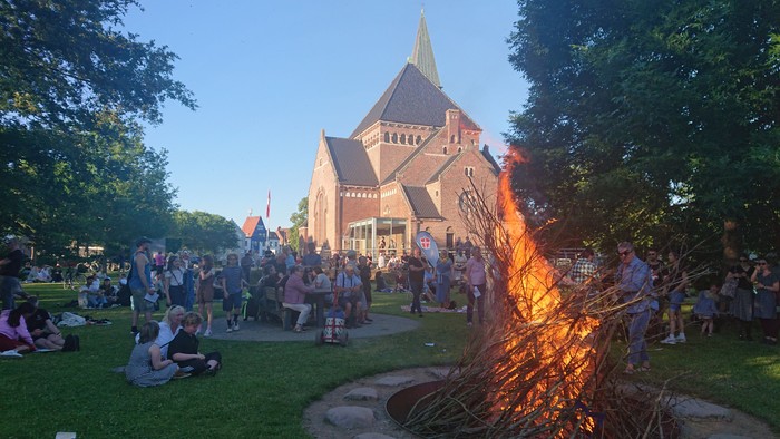 En gruppe af mennesker sidder på græs og ser en stor bålstokke ved en kirke.