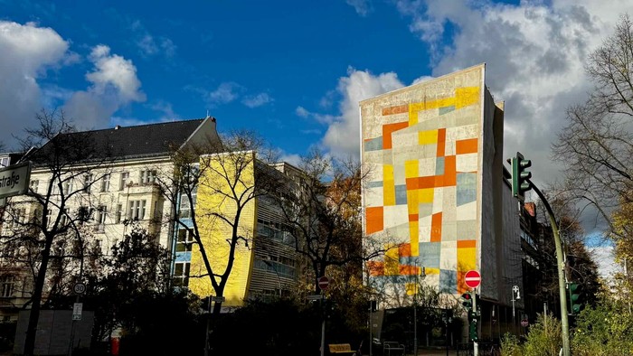 Städtische Szene mit einem farbenfrohen geometrischen Wandgemälde an einem modernen Gebäude neben traditioneller Architektur unter blauem Himmel.