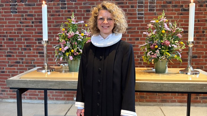 Kvinde i svart kapelletøj foran mur med blomster og lysstake