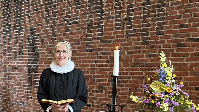 En kvinde i svart kapittel holder en bog og står foran en mur af røde mursten.