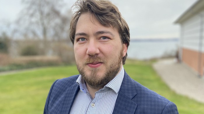 En mand i jakkesæt står udendørs på et græsklædt område med en bygning og vand i baggrunden.