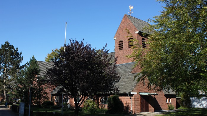 Eine Backsteinkirche mit hohem Turm und Wetterhahn, umgeben von üppigen grünen Bäumen unter einem klaren blauen Himmel.